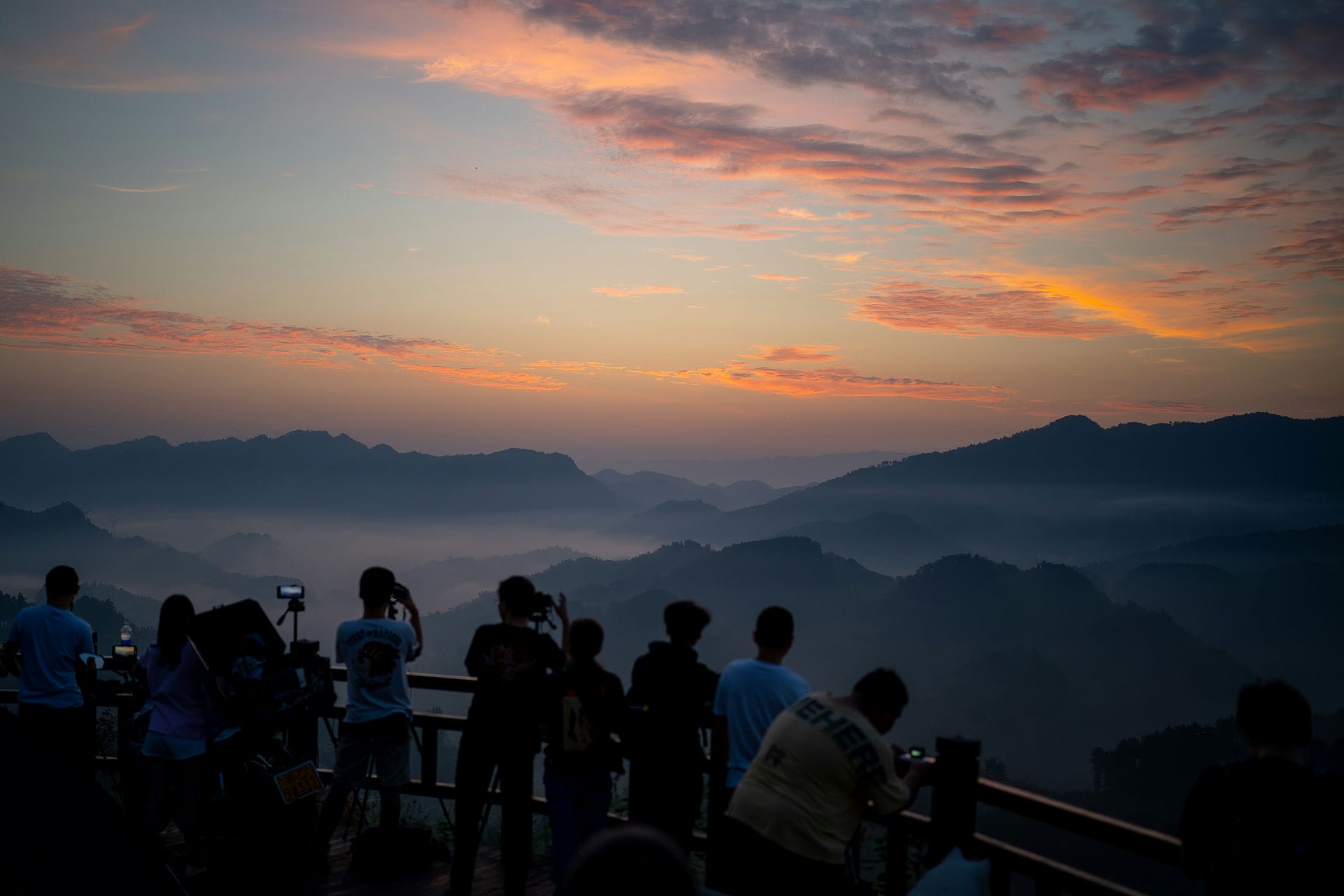 周家山大桥日出时,观景平台上的人影面对被晨光染暖的天空,远处山脊与山谷薄雾层层退远。